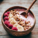 A bright, overhead food photography shot of a creamy raspberry smoothie bowl topped with fresh raspberries, banana slices, and crunchy granola. A wooden spoon rests inside the bowl, angled as if ready to scoop. Natural daylight from the side highlights the textures of the fruit, granola, and creamy base. Shot on a rustic light wooden table for a fresh, wholesome feel. First Image